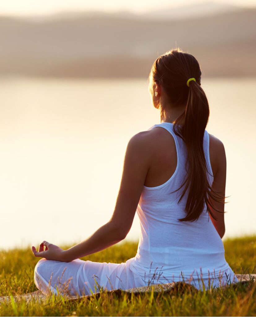 a woman sitting in the grass meditating while facing a tranquil lake